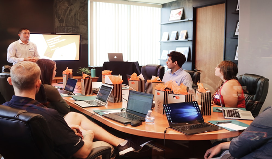 give employees at a boardroom table with laptops, listening to a speaker standing at the head of the table with a projector.