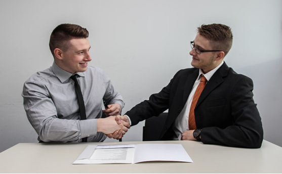 two men at a desk shaking hands over a contract file.