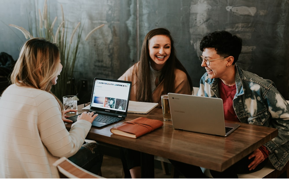 3 employees laughing at a table together with a couple laptops and some notebooks.