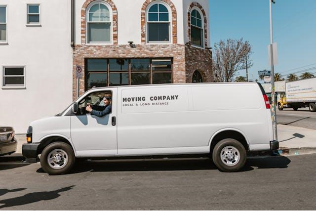 a white moving van in the street with the driver sticking his head out the window to look back.